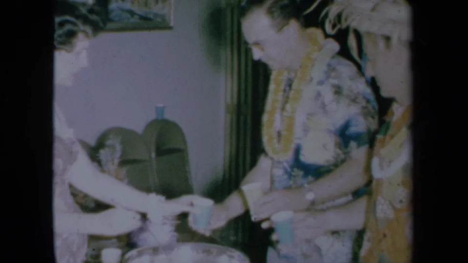 An adult man in a Hawaiian shirt and lei accepts a drink beside the punch bowl at an indoor party.