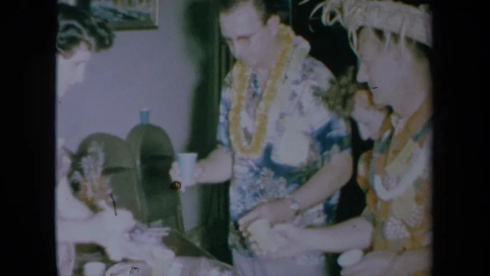 1961 California home-movie frame showing adults in leis and aloha-style clothing around a party table in a private interior.