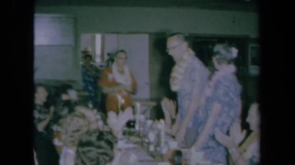 Wide 8mm home-movie view of a crowded 1961 California party room with seated guests, standing adults, leis, and table settings.