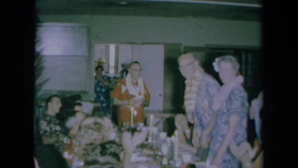 1961 California home-movie frame showing adults in leis and patterned shirts standing beside seated guests at an indoor party.