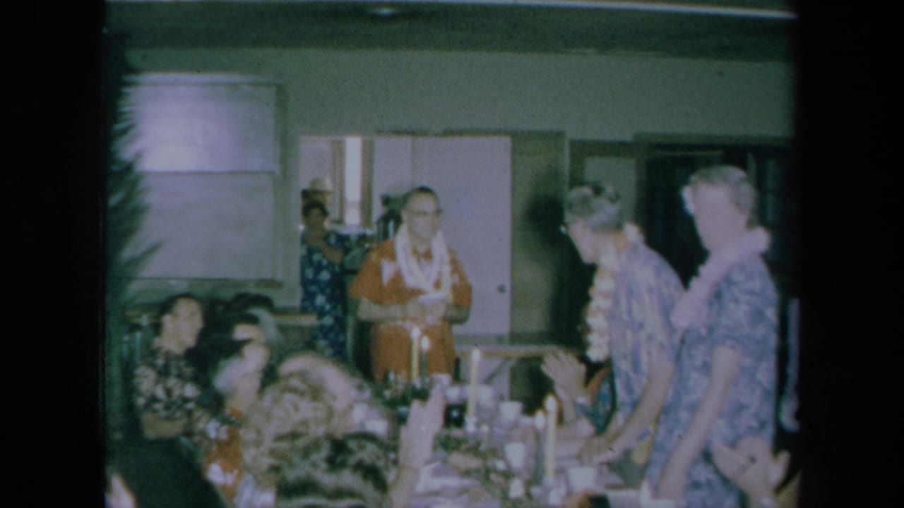 1961 California home-movie frame of a Hawaiian-themed indoor meal with guests seated at a long table and a man standing to speak.