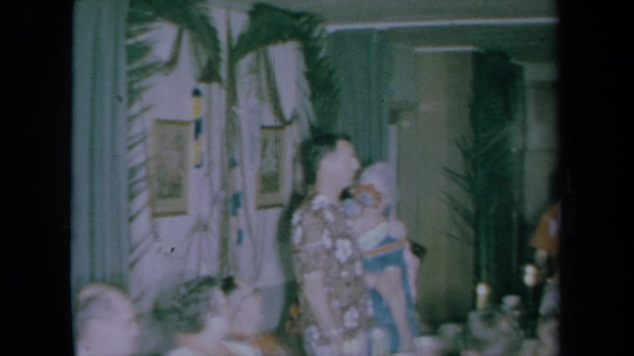 A man in a patterned shirt stands near wall decorations while holding or greeting a guest beside the dining table.