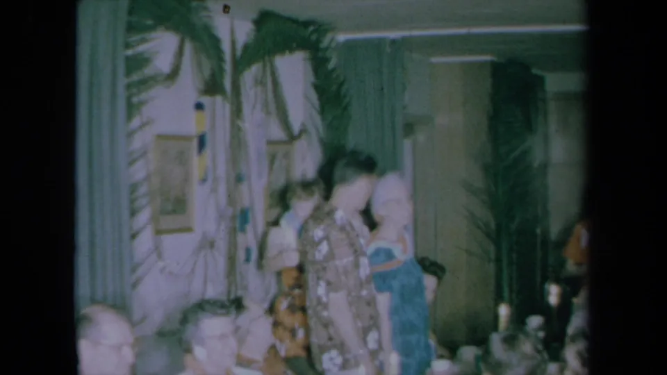 1961 California home-movie frame of party guests in aloha-style clothing embracing or talking beside a candlelit dining table.