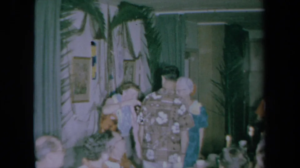 1961 California home-movie frame of a patterned-shirt guest seen from behind during a Hawaiian-themed indoor family celebration.