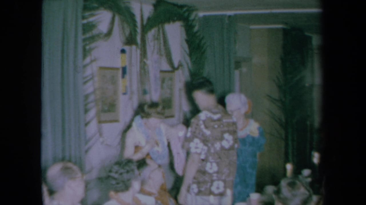 1961 California home-movie frame showing party guests indoors around a lei exchange, with palm fronds and curtains behind them.