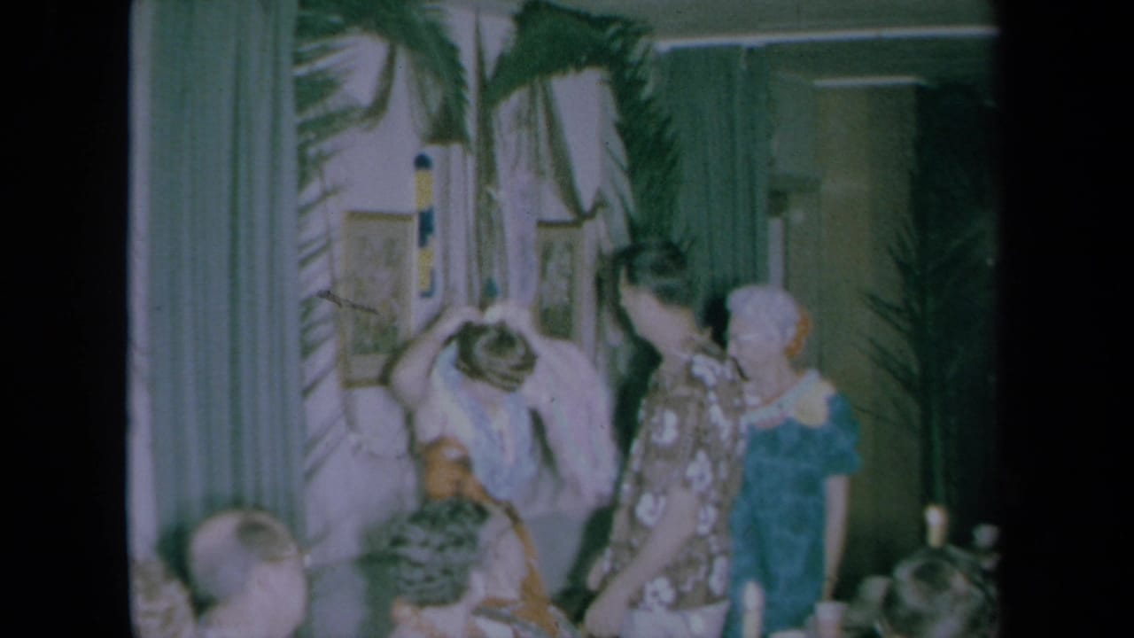 A woman lifts or removes a lei while standing near a man in a floral shirt and other guests.