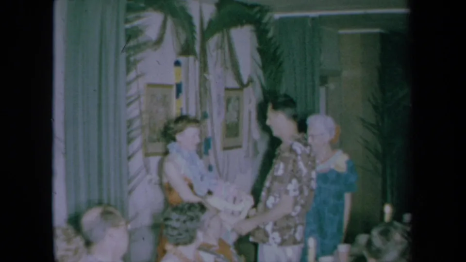 1961 California home-movie still with a woman and man facing each other during a lei presentation in a tropical-themed room.