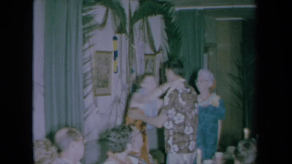 1961 California party frame showing a lei being placed or adjusted near a man in a floral shirt while guests observe.