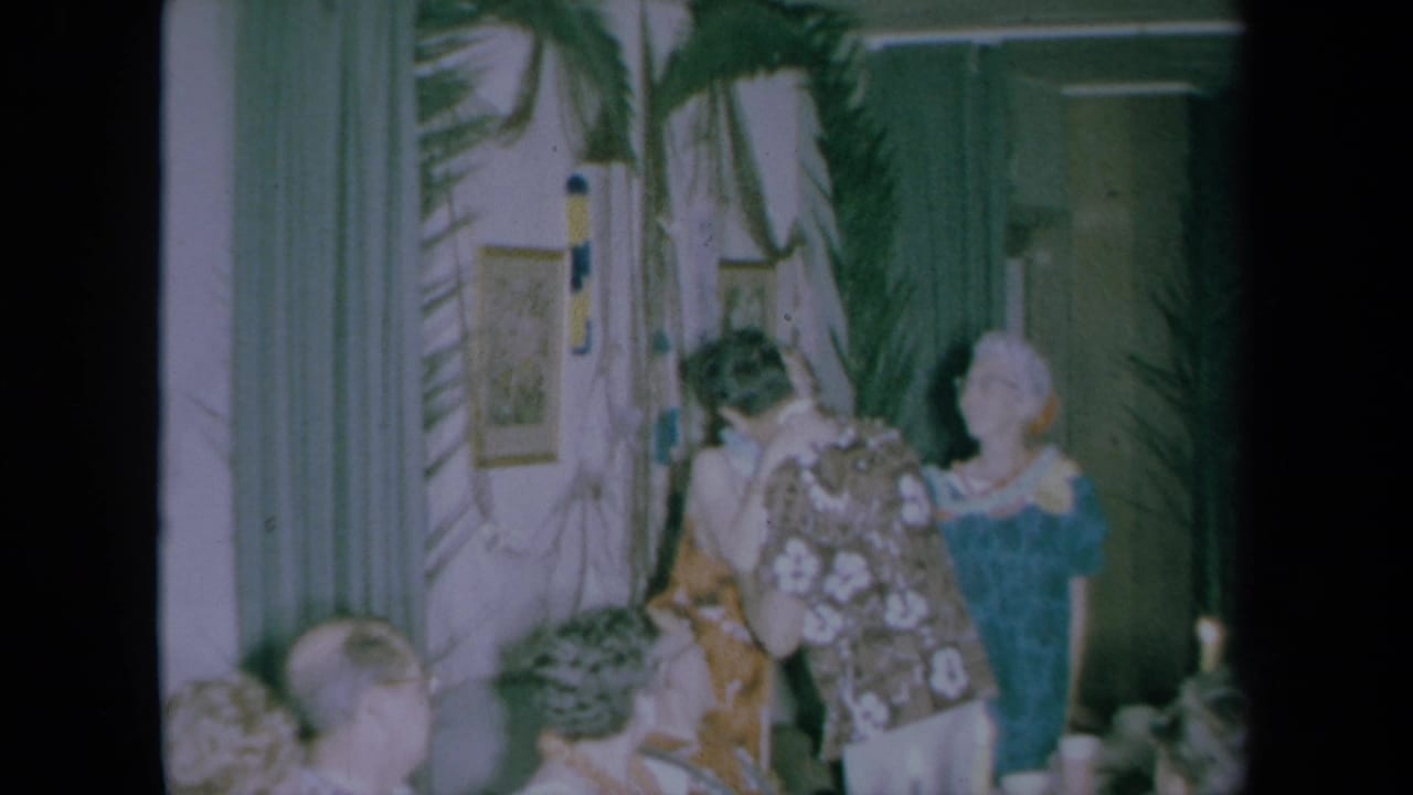 Guests gather in a decorated room as a woman in a floral dress greets people near palm fronds.