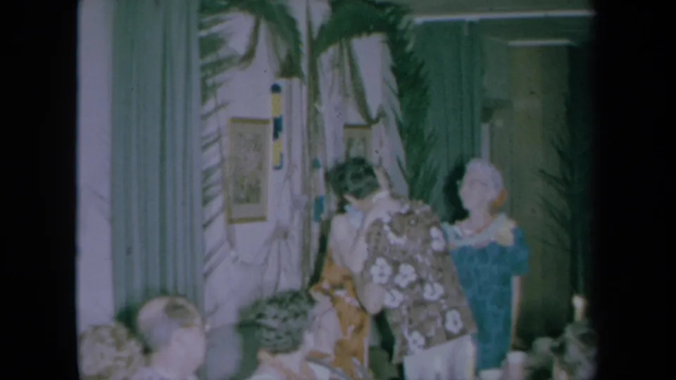 In 1961 California home-movie footage, party guests stand and sit in a decorated room with palm fronds, curtains, and tropical clothing.
