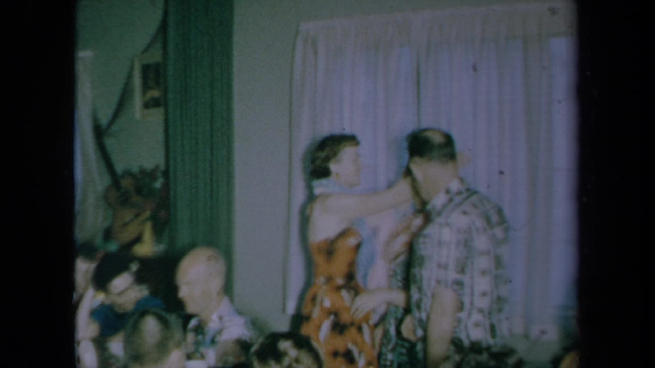 A woman in a floral dress greets a man in a patterned shirt beside pale curtains.