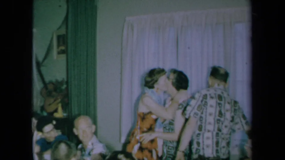 At the indoor California party, a woman kisses a guest while another man in a patterned shirt stands nearby against pale curtains.