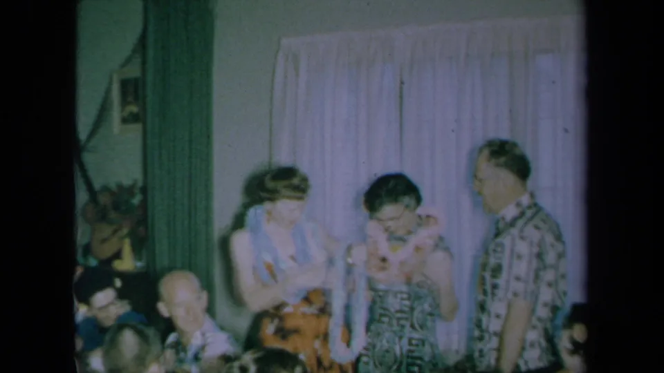 The 1961 home movie shows the woman and guest after the greeting, with a lei visible and another man observing in the decorated room.