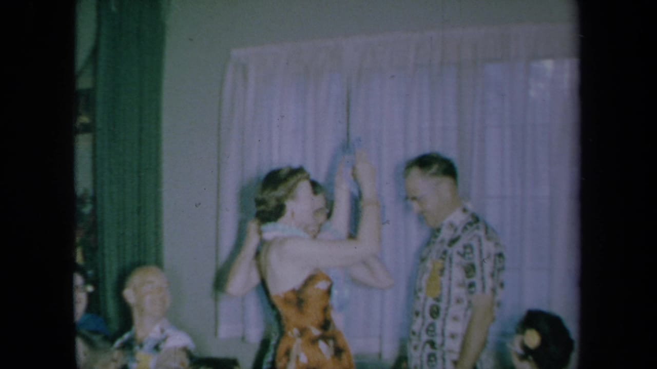Woman raises a flower lei beside a man in a tropical shirt as seated guests watch indoors.