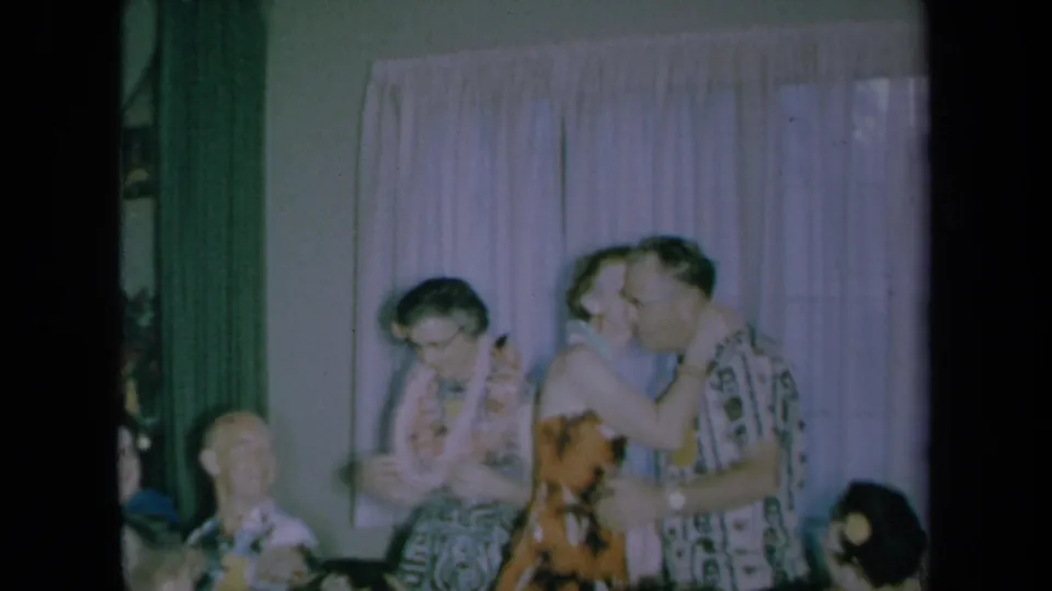 A woman kisses a man after placing a lei around his neck during a 1961 California indoor home gathering.