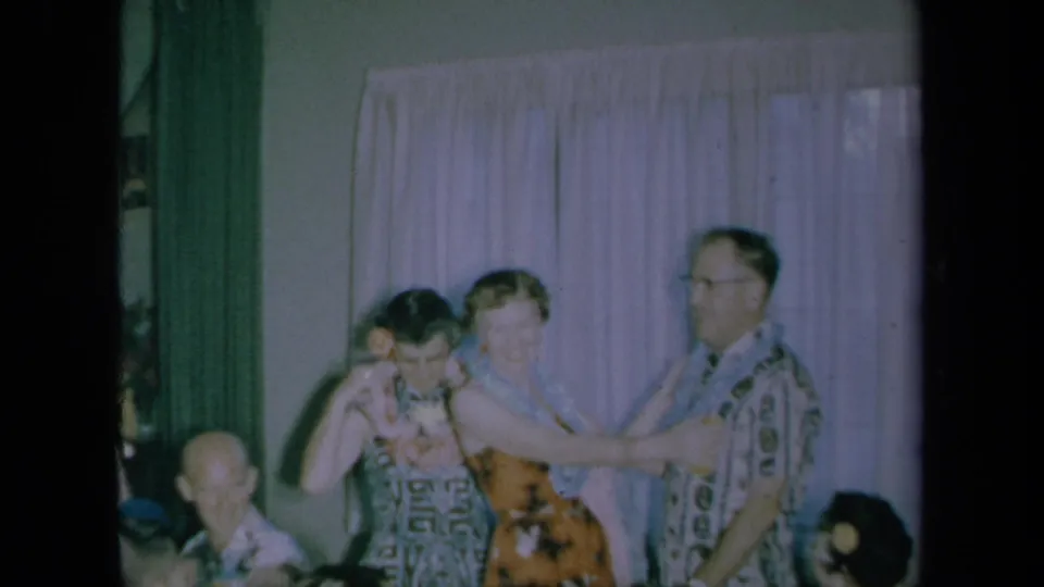 The woman and man stand before a curtain as the lei presentation continues in a 1961 California home-movie party scene.