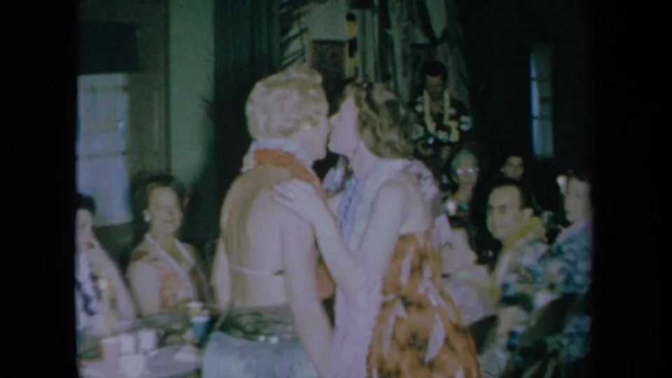 A 1961 California home-movie frame shows a kiss at a crowded indoor party with leis, tropical shirts, and seated guests.