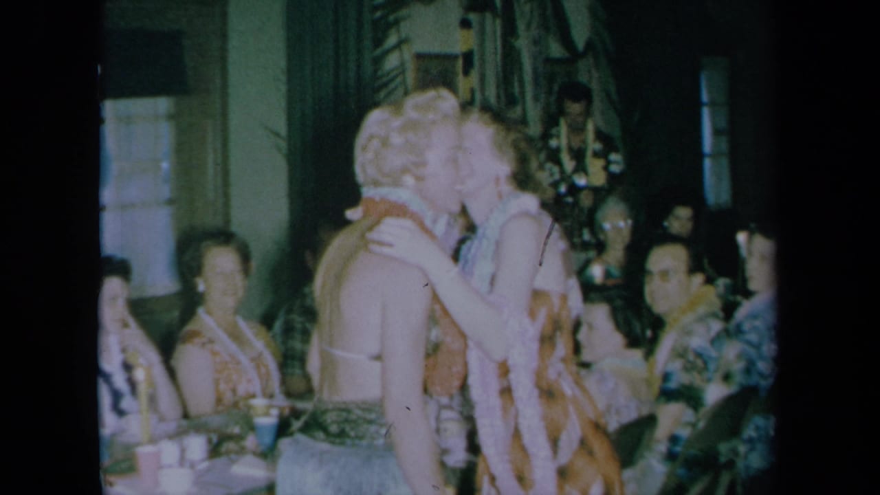 Two women wearing floral leis kiss during an indoor celebration while guests sit nearby.