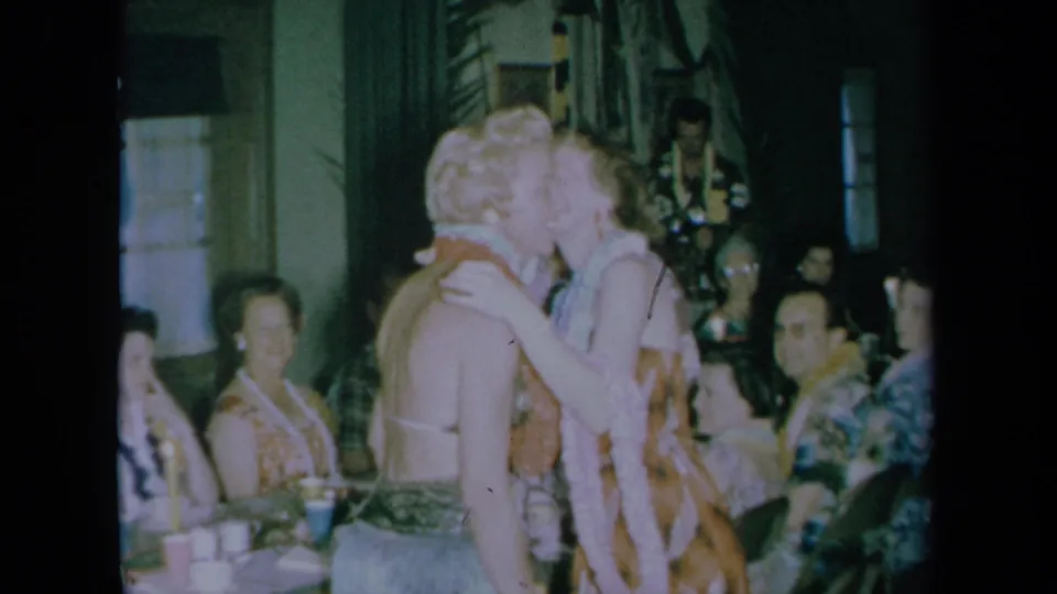 In a 1961 California home-movie frame, two women in leis kiss at a dim indoor wedding celebration with seated guests behind them.
