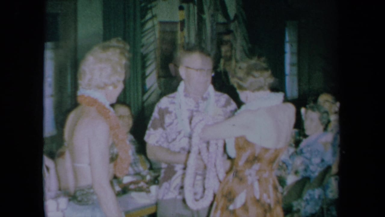 A woman places or adjusts a lei on a man as another woman watches in a crowded room.