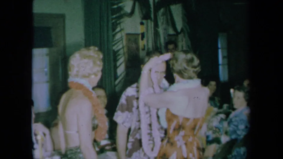 A woman with her back to the camera raises her arms near another lei-wearing woman and a man amid seated guests in the 1961 celebration.