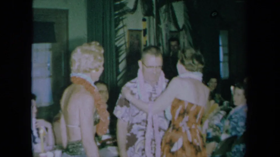 A woman in a patterned dress adjusts a man's lei as another woman wearing a lei stands nearby during the 1961 California indoor wedding celebration.