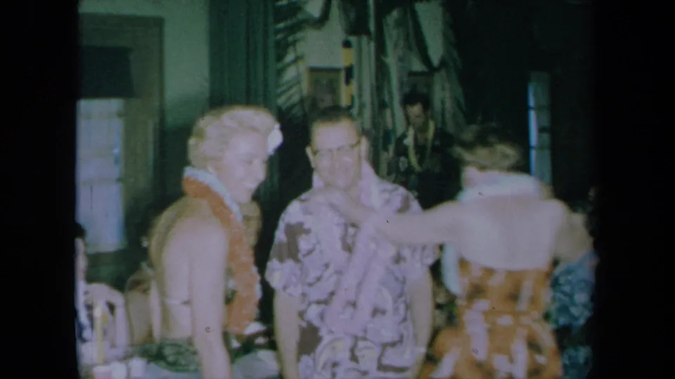 A woman in a lei smiles beside adults in floral clothing during the 1961 California party footage.