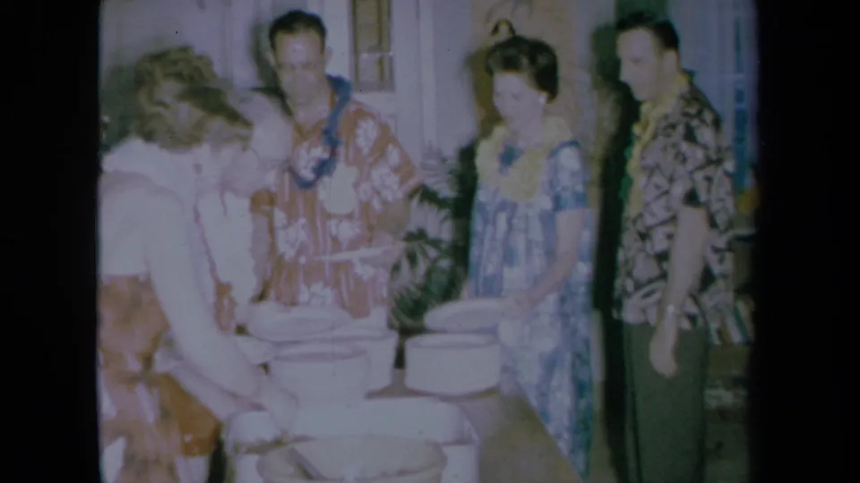 Guests wearing leis stand by a buffet table with white bowls in the 1961 California home movie.