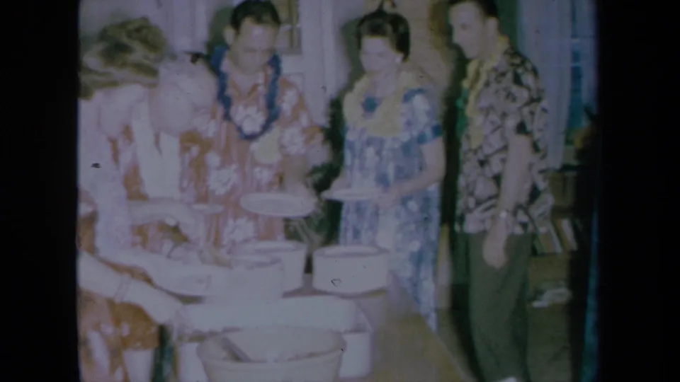 A woman serves from white bowls while lei-wearing guests wait in the 1961 California party scene.