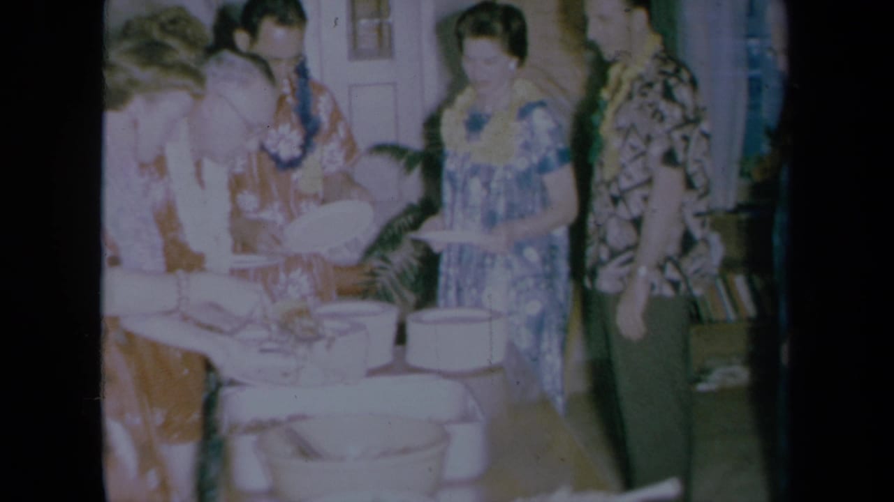Adults in leis and patterned clothing gather around a buffet table with plates and bowls.