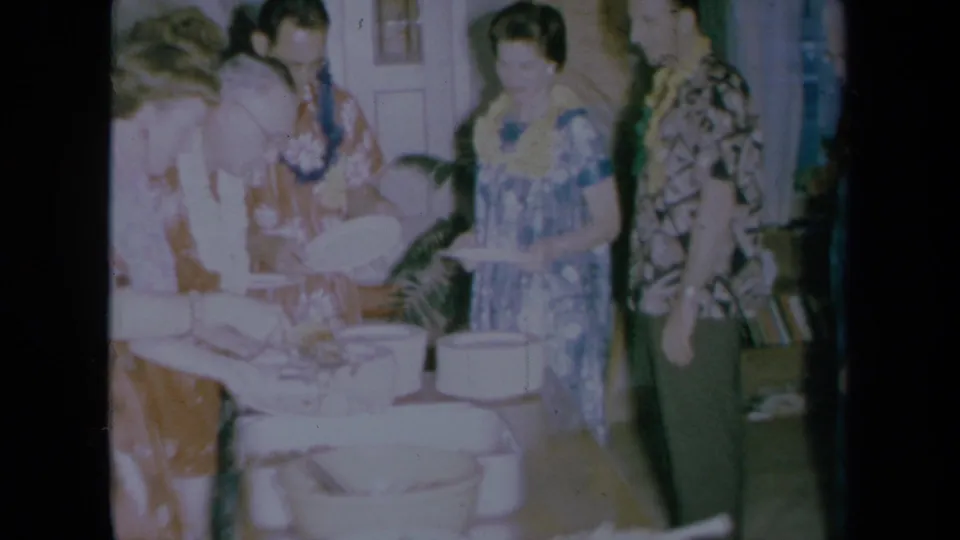 1961 California home-movie frame showing adults at a Hawaiian-themed buffet with leis, plates, and serving bowls inside a private room.