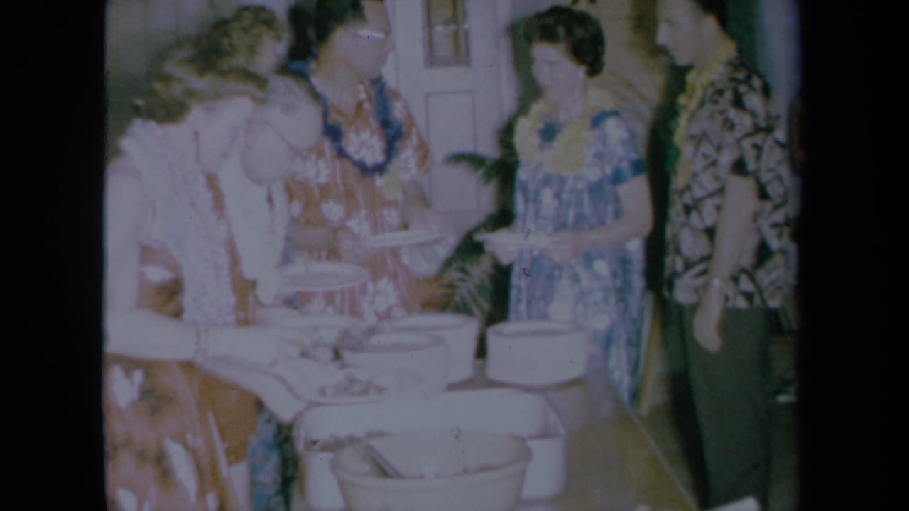 Adults in aloha-style clothing and leis stand by buffet dishes in a private California party setting, filmed on soft 8mm color stock.