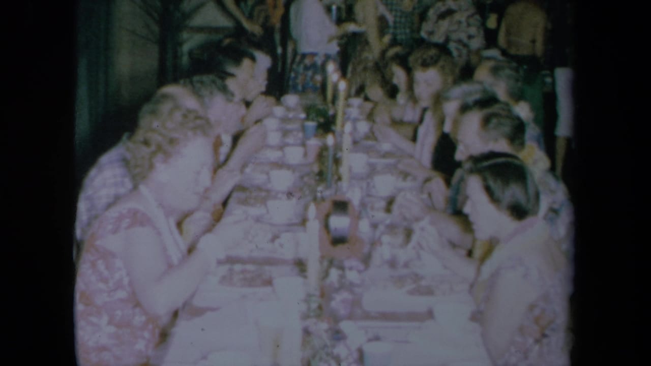 1961 California home-movie frame showing adults gathered along a long dining table during a meal.