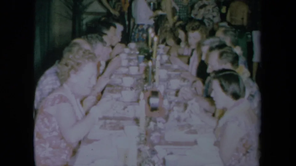 1961 California home-movie frame showing adults gathered along a long dining table during a meal.