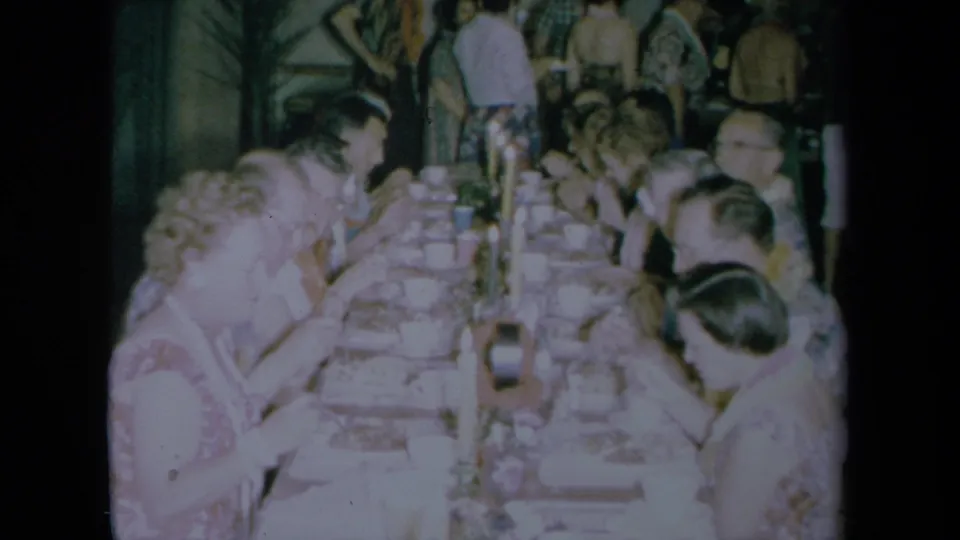 Adults in period clothing eat and talk around a long rectangular table in 1961 California footage.