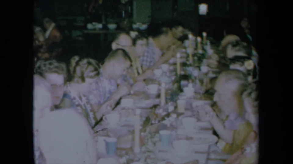 Home-movie view of adults eating at a long holiday table in a dim California private interior, with 8mm grain and soft color.