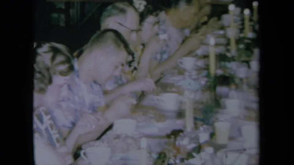 Closer 1961 California home-movie frame of adults eating beside a long candlelit dining table with bowls, cups, and plates.
