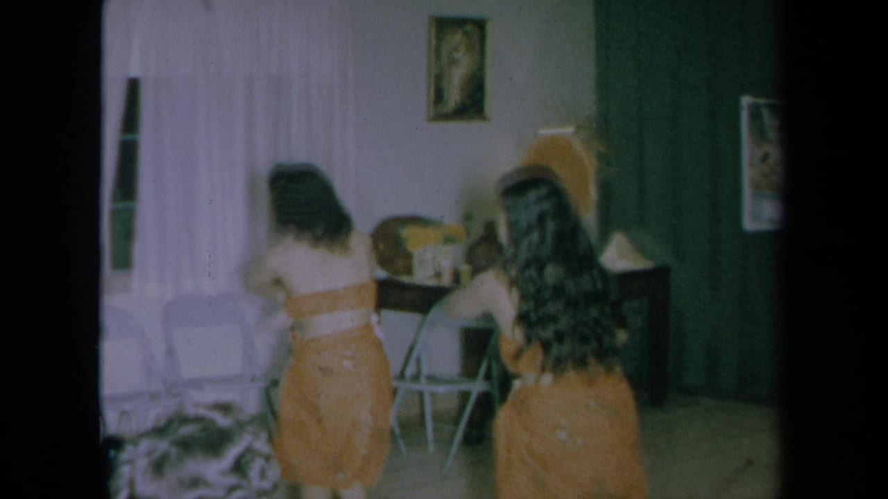 Young women in orange dresses stand near furniture in a curtained room.
