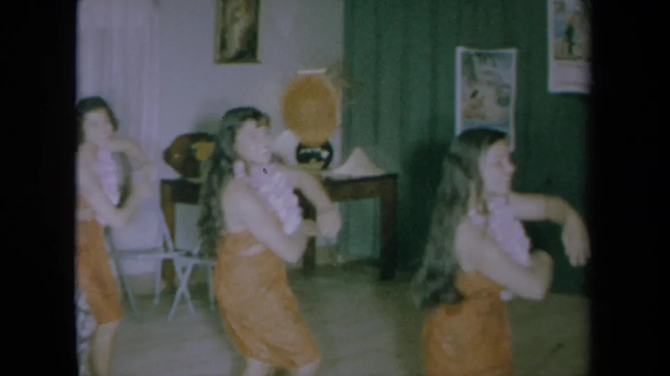 1961 California home-movie frame of three young women in orange Hawaiian-style dresses performing a dance in a private interior.