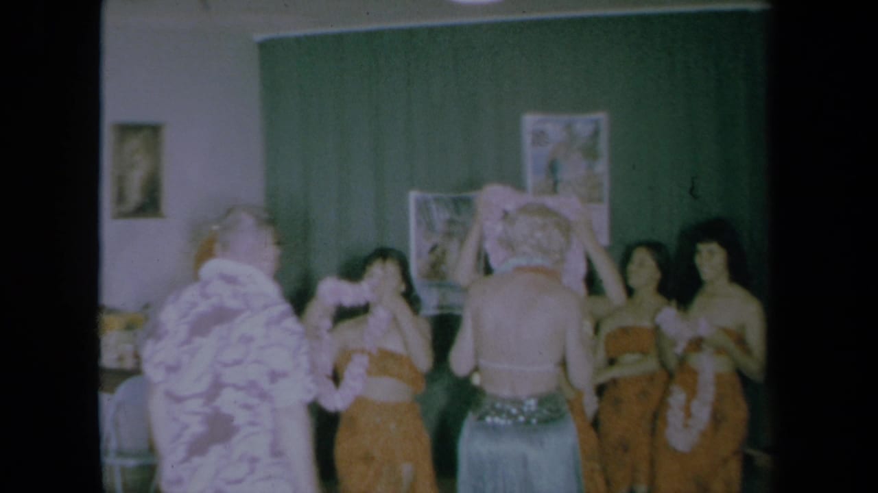 1961 California home-movie frame showing hula-costumed women welcoming a man and woman with leis inside a private room.