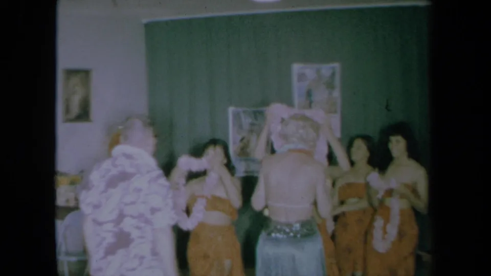 1961 California home-movie frame showing hula-costumed women welcoming a man and woman with leis inside a private room.