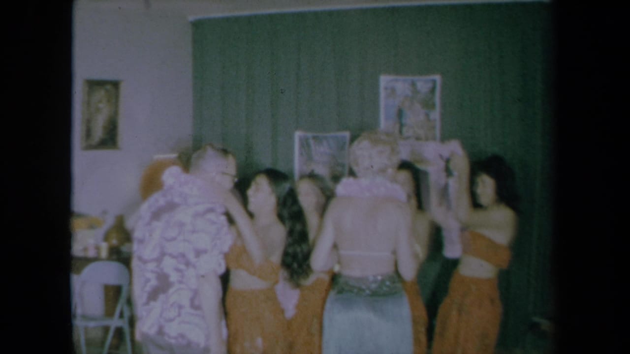 A group surrounds two guests as leis are raised near their necks.