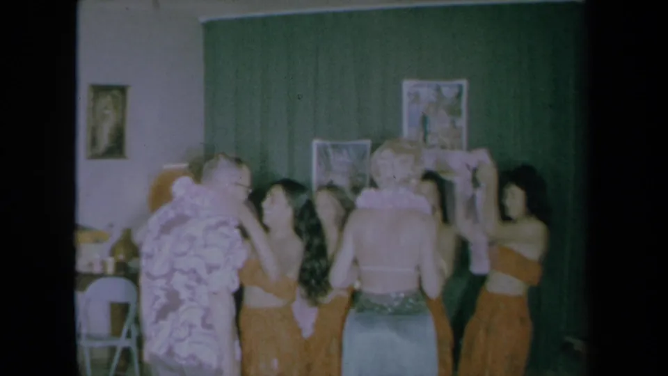 1961 California home-movie frame with women in orange hula-style outfits placing leis on two guests in a private interior.