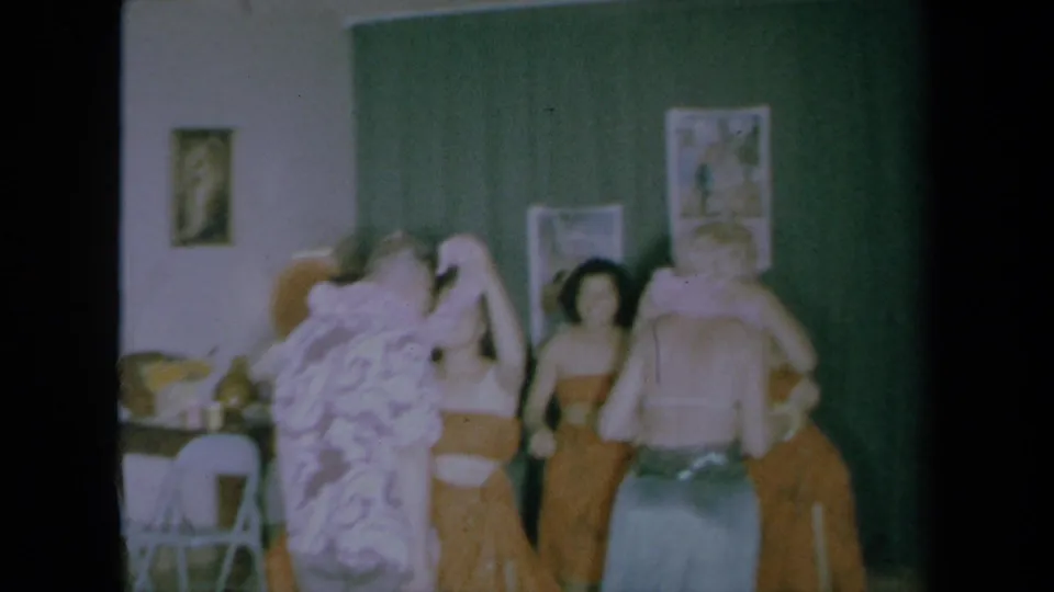 1961 California home-movie frame showing a close group during a Hawaiian-themed lei welcome in a room with curtains and wall art.