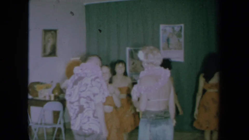 1961 California home-movie frame with guests and hula-costumed women near green curtains as the lei welcome winds down.