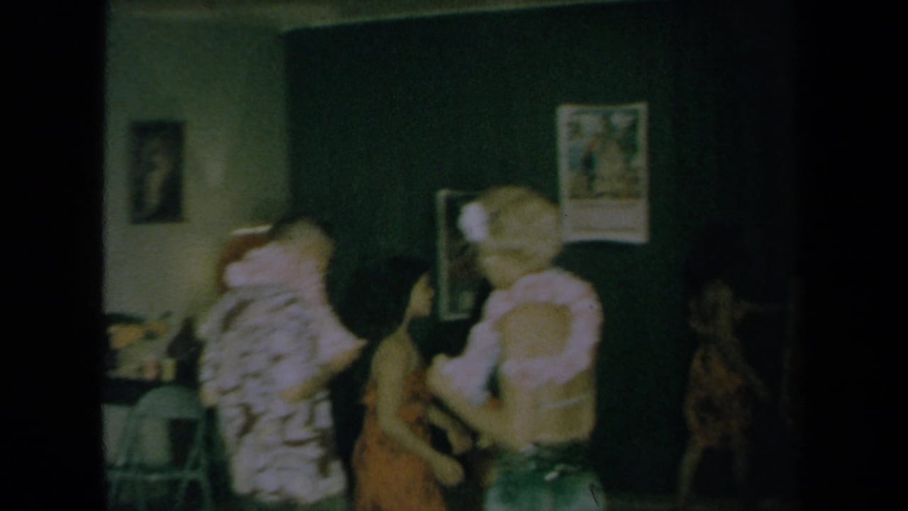 1961 California home-movie frame showing an indoor Hawaiian-inspired dance scene with blurred dancers and onlookers near wall posters.