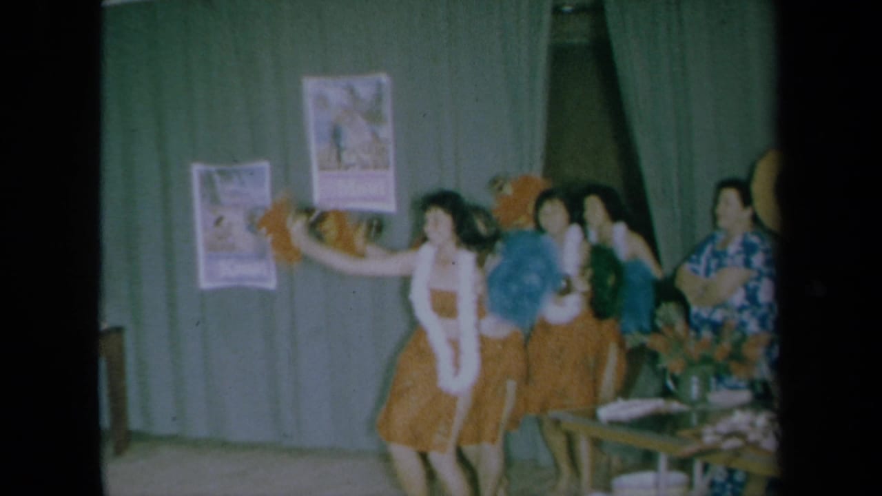 Women in grass skirts and leis perform in front of green curtains.