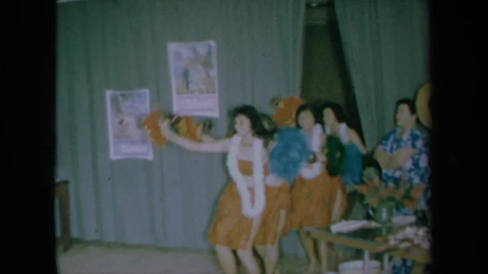1961 California frame of women performing a Hawaiian-inspired dance with leis, grass skirts, and colorful props before green curtains.