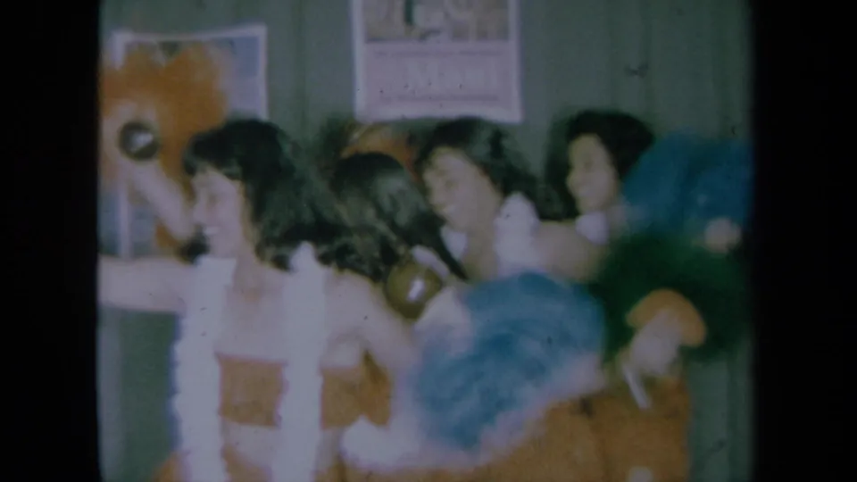 1961 California frame showing women continuing a Hawaiian-inspired dance, with leis, grass skirts, props, posters, and green curtains visible.