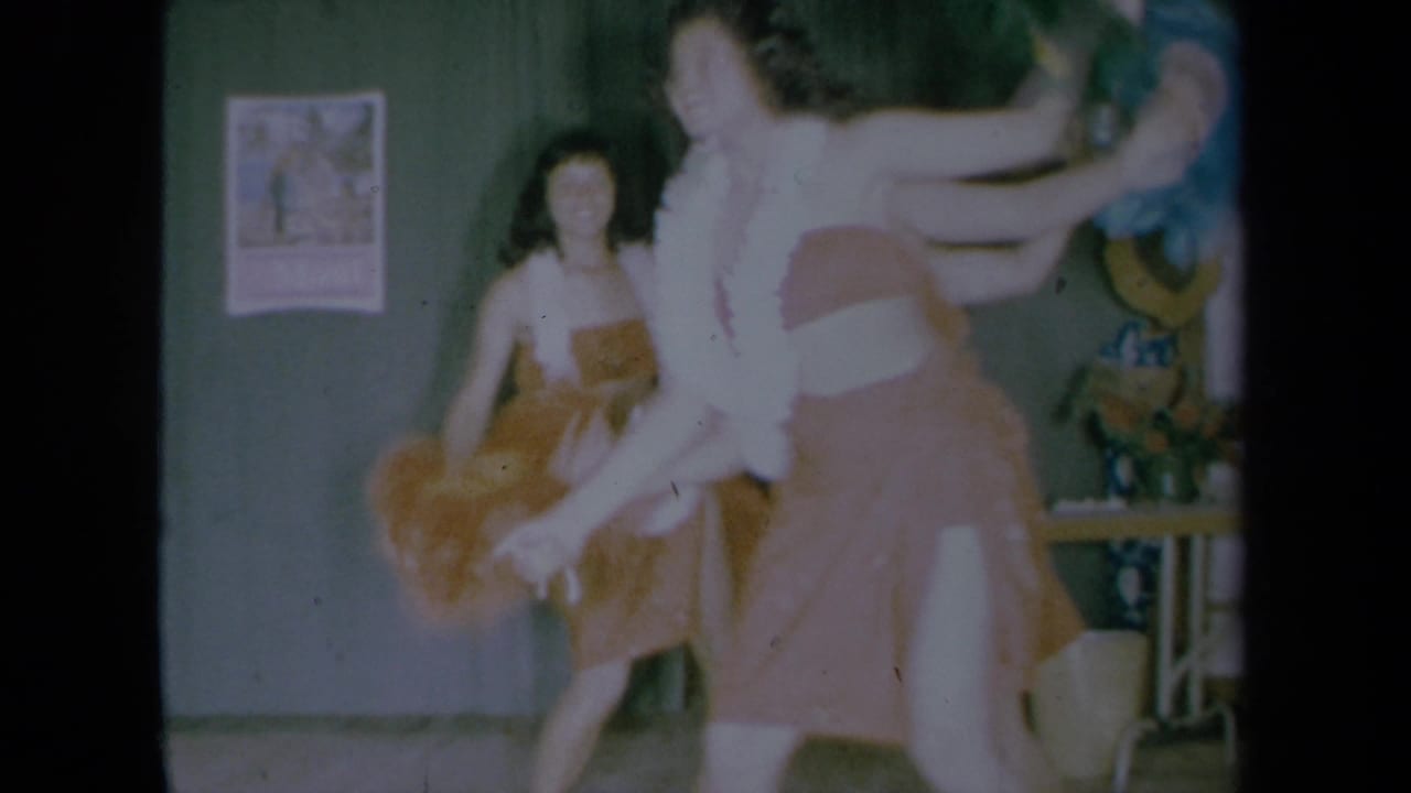 Two young women dance in a California home interior, one in a pink dress with a white feather boa and another in a brown dress.